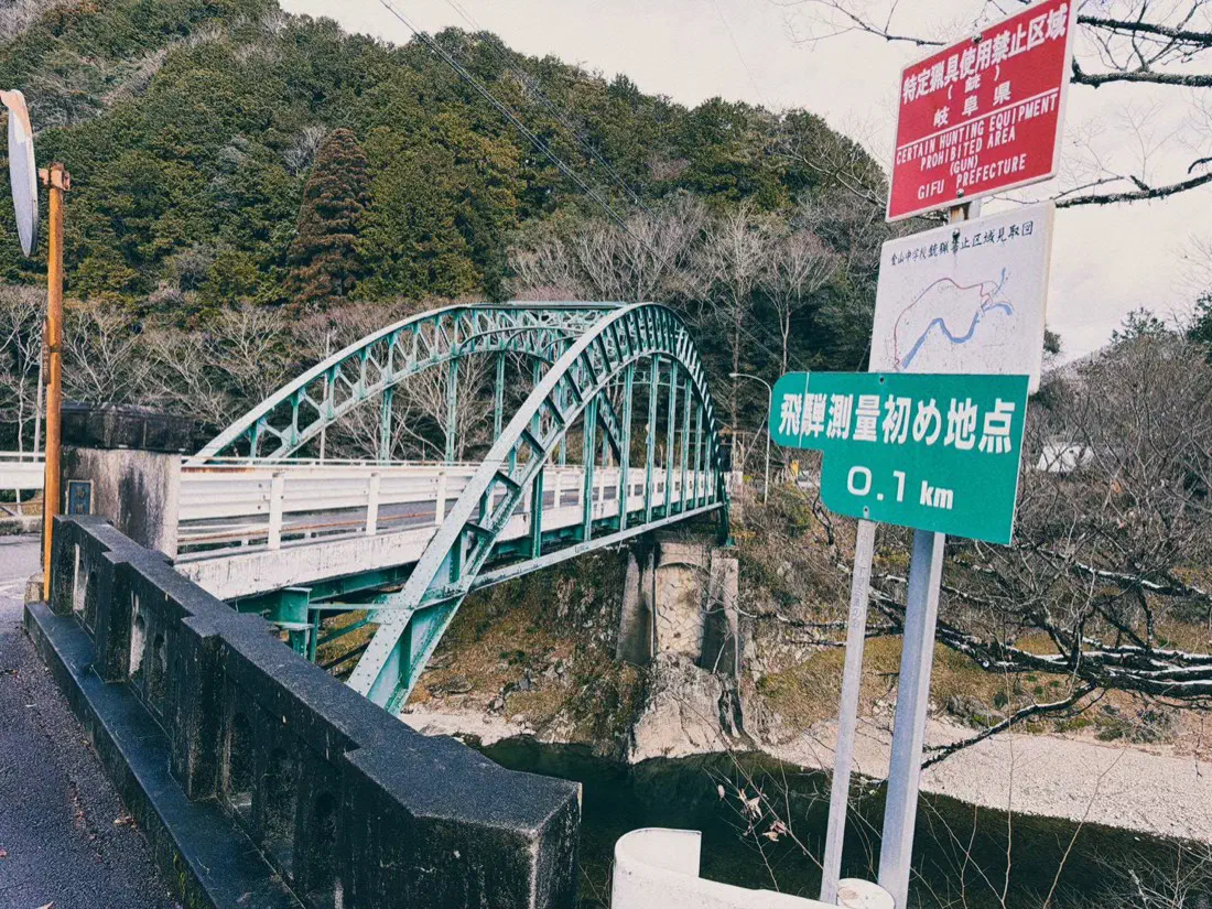 川にかかる緑のアーチ橋と、「飛騨測量初め地点」の標識